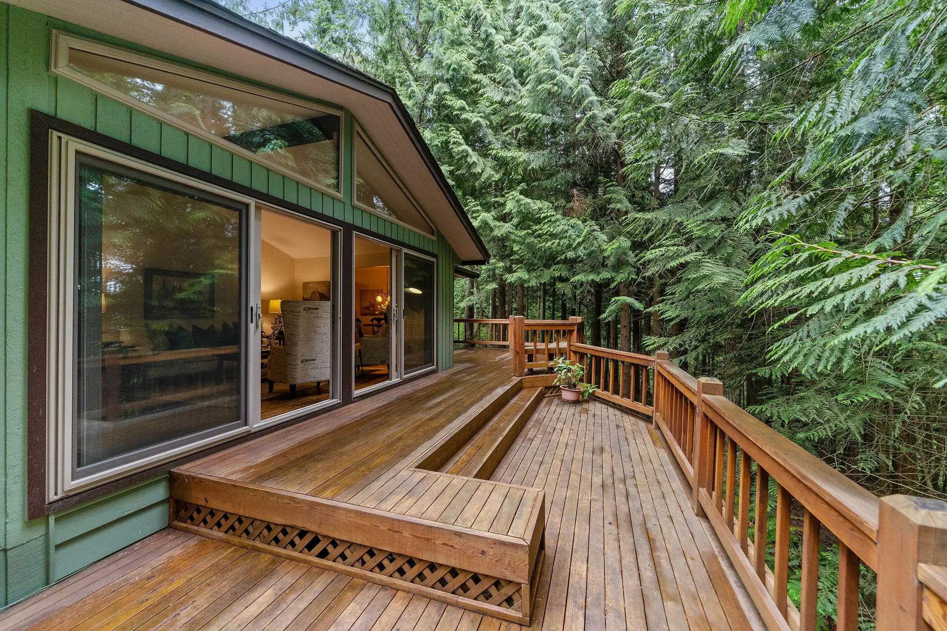 Stock photo: a wraparound wood deck with a built-in bench and wood railing beside a green house, surrounded by evergreen trees